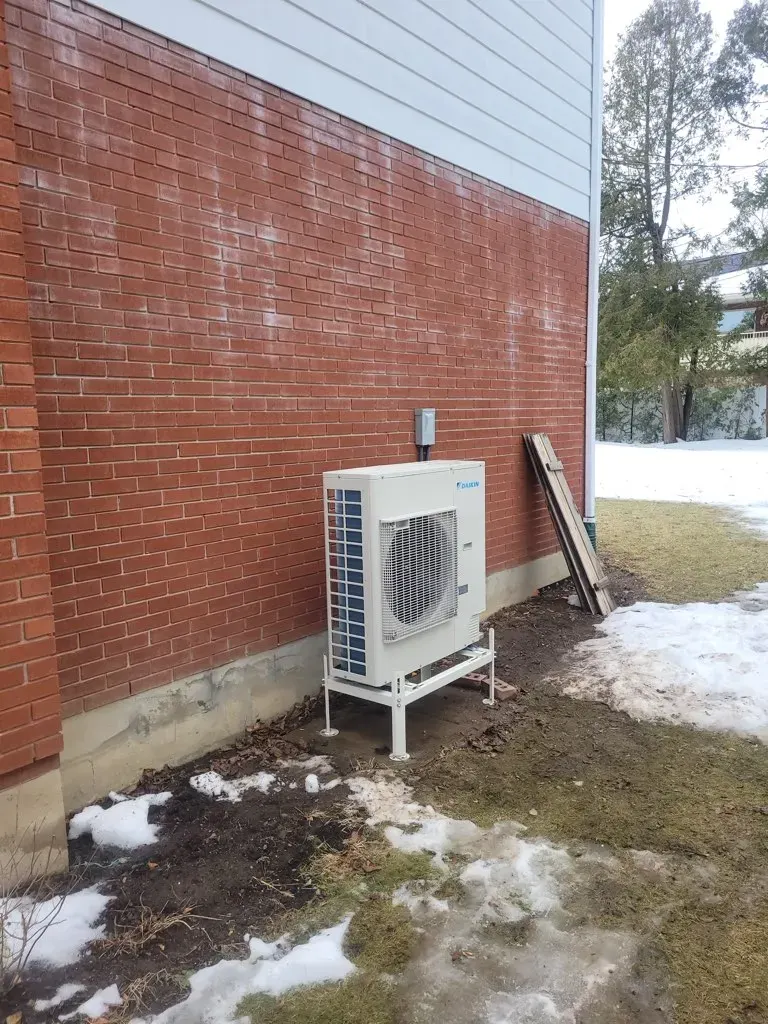 A heat pump condenser mounted on a wall bracket beside a brick home in winter